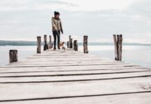 Frau in herbstlicher Kleidung geht mit einem kleinen Hund am See spazieren