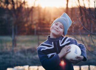 ein Junge mit Trainingskleidung und Mütze strahlt voller Selbstvertrauen und hält einen Fussball in seinen Händen
