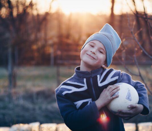 ein Junge mit Trainingskleidung und Mütze strahlt voller Selbstvertrauen und hält einen Fussball in seinen Händen